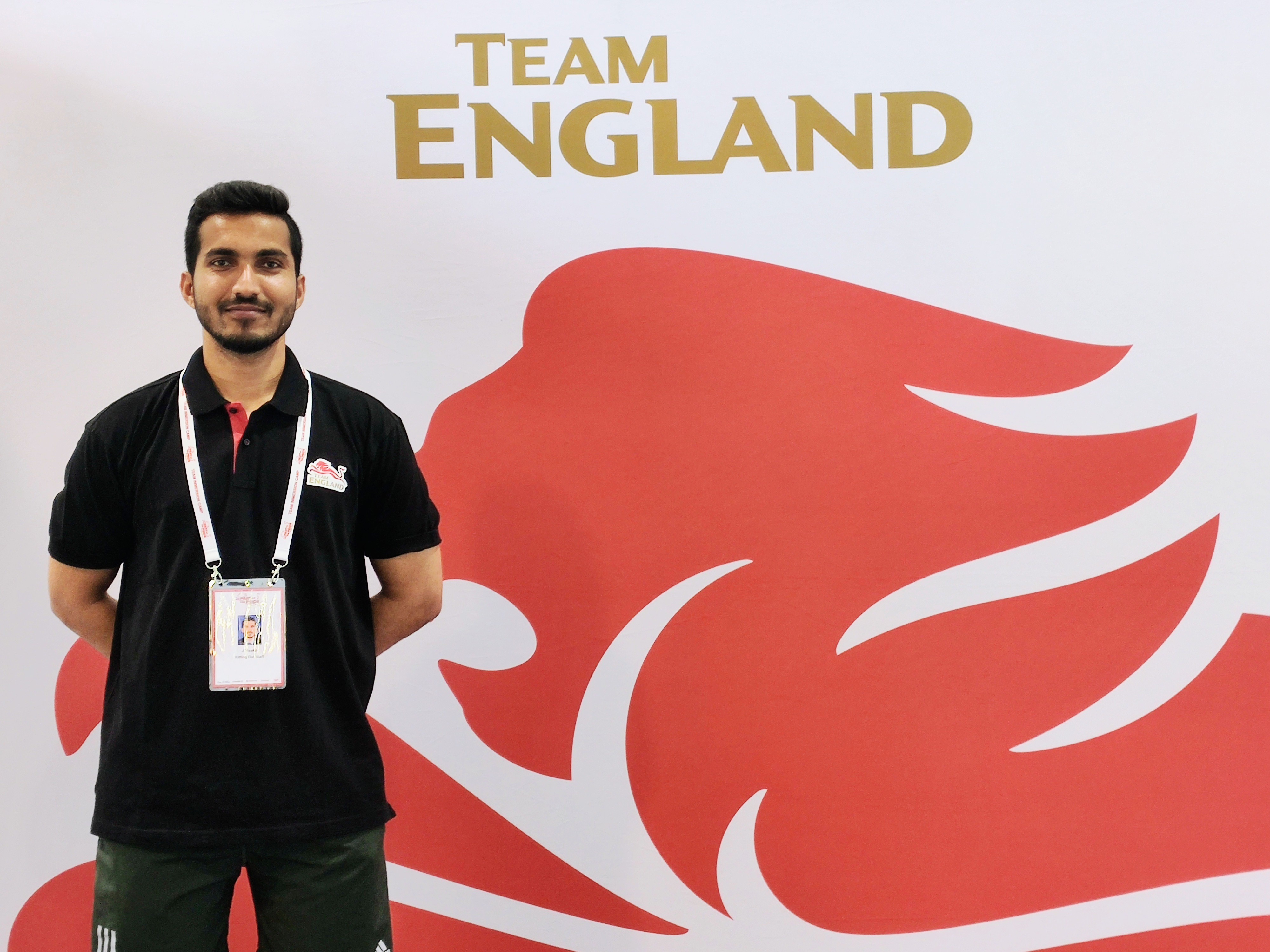 Visakh standing in front of a Team England background whilst volunteering for the Commonwealth Kitting Out team. 